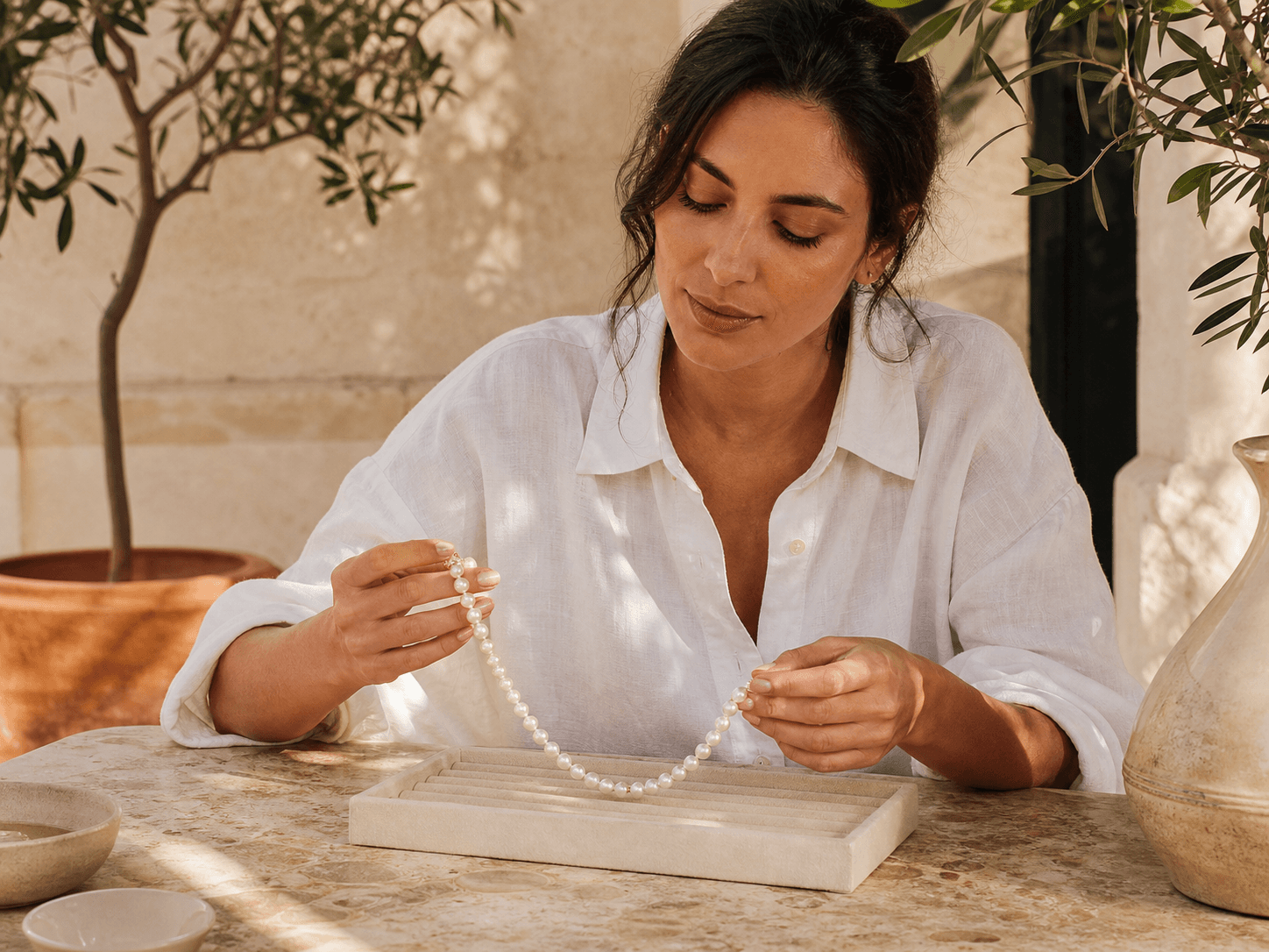 Model examining a pearl necklace at a sunlit limestone table